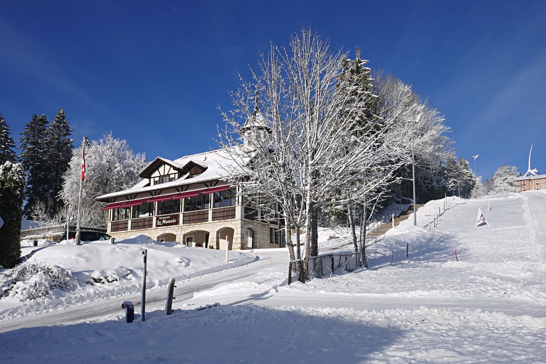 Le Manoir et le haut de Mont-Soleil sous la neige et le soleil, une éolienne au loin.